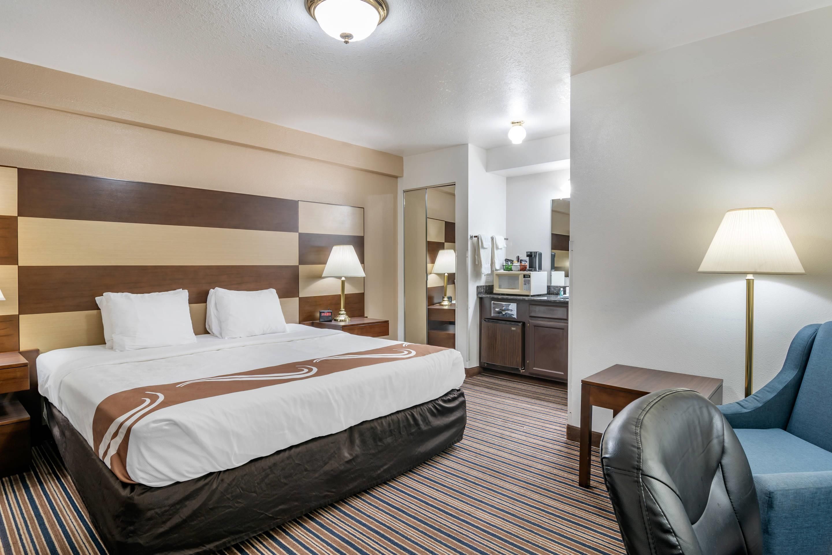 A hotel room with a large bed, bedside tables, a desk, a chair, a kitchenette, and a mirror.
