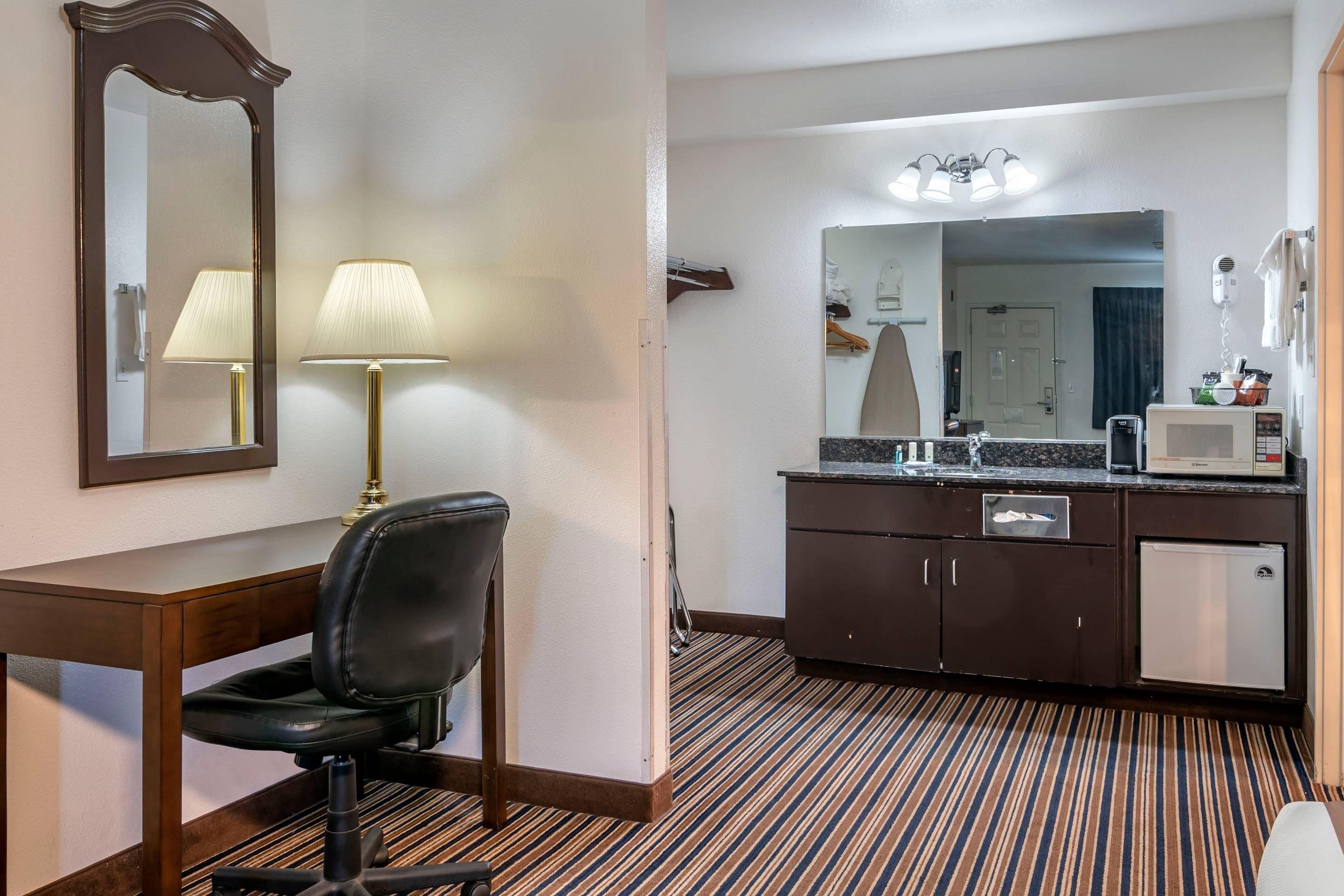 Habitación de hotel con escritorio, silla, lámpara, espejo grande, tocador con lavamanos y microondas.