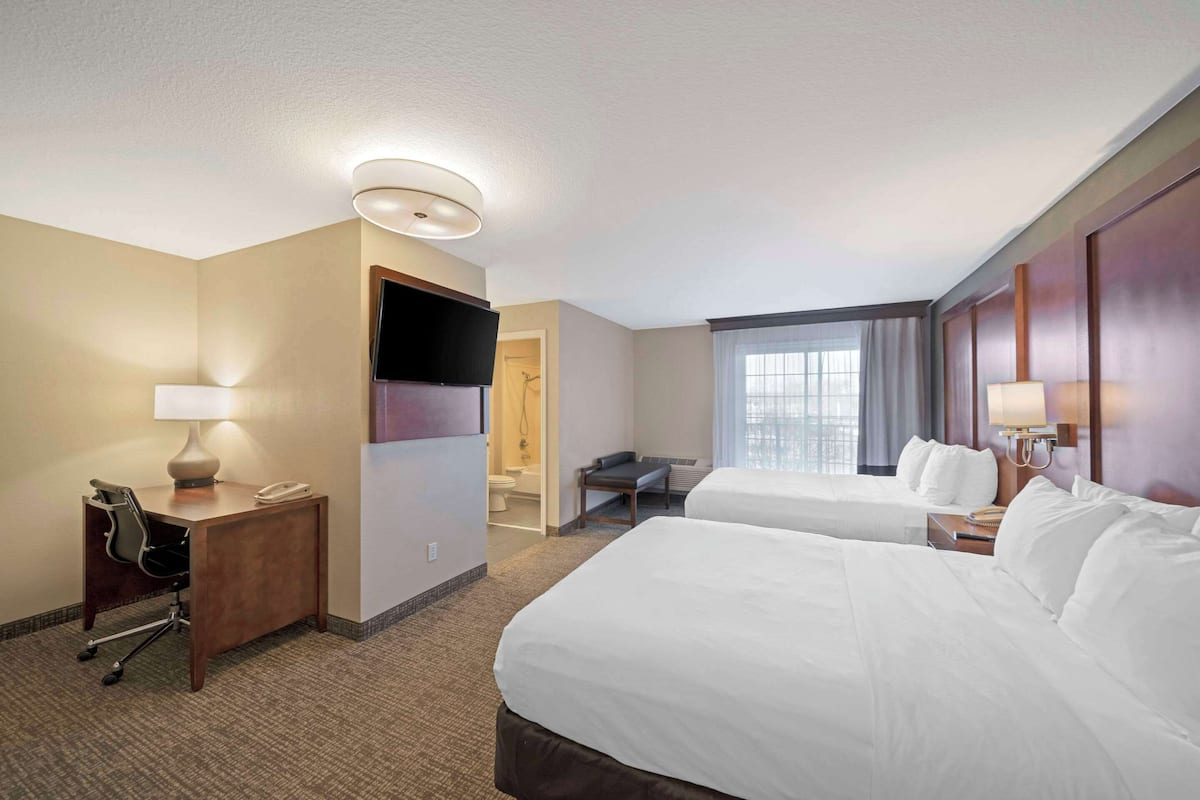 A hotel room with two beds, a desk with a lamp, a TV, and a bathroom visible through an open door.