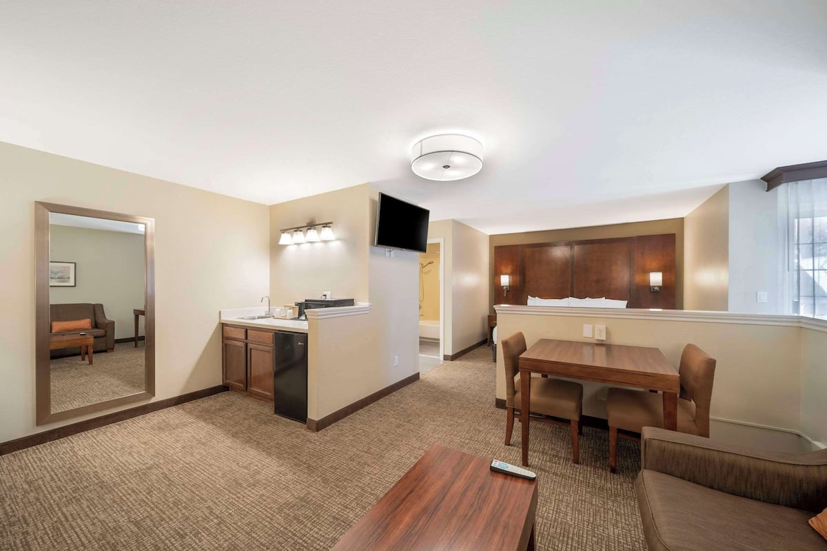 A hotel room with a kitchenette, a dining area with a table and chairs, a sofa, a TV mounted on the wall, and a large mirror reflecting the room.