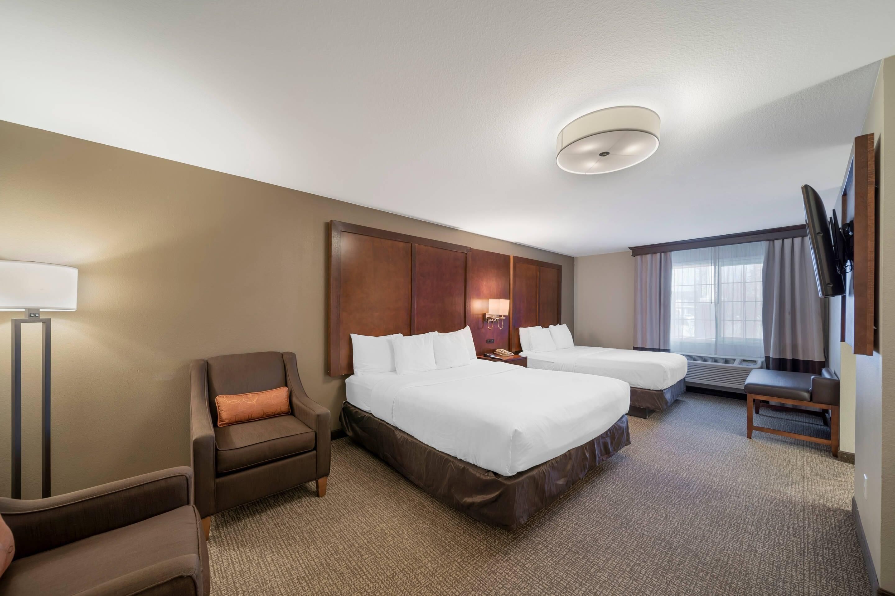 A hotel room with two beds, a chair, a desk, a lamp, and a TV.