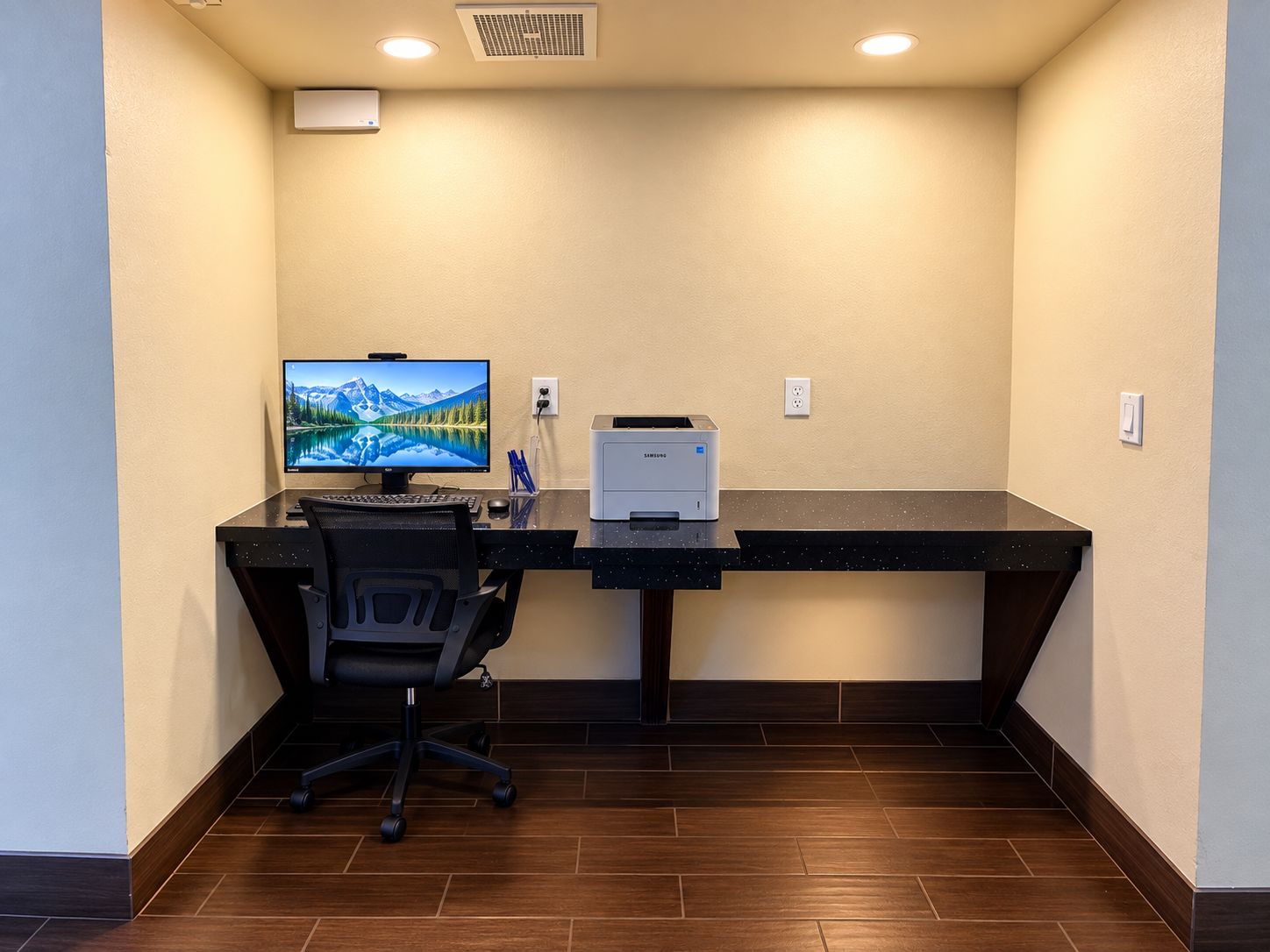 A hotel room with a desk, computer, printer, and a chair.