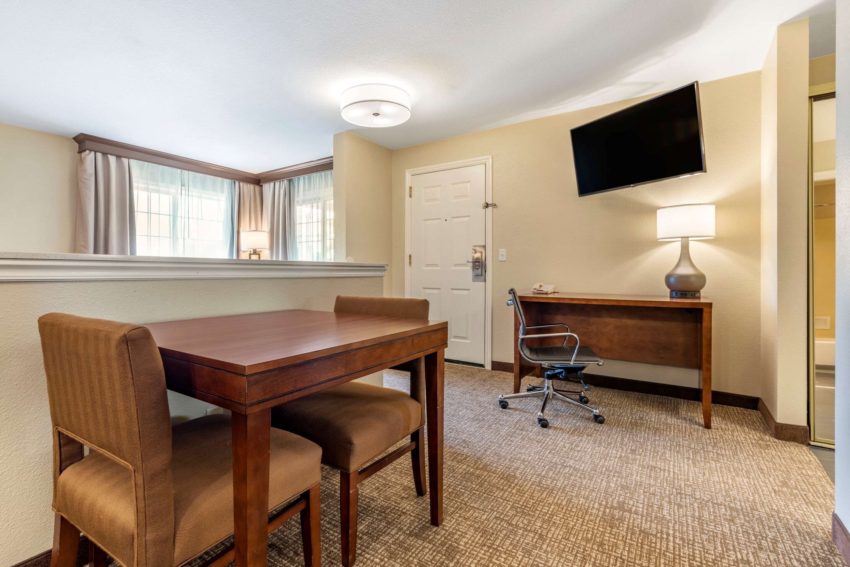 Une chambre d’hôtel comprenant une table à manger en bois, deux chaises rembourrées, un bureau avec une chaise, un téléviseur à écran plat fixé au mur et une lampe sur le bureau.