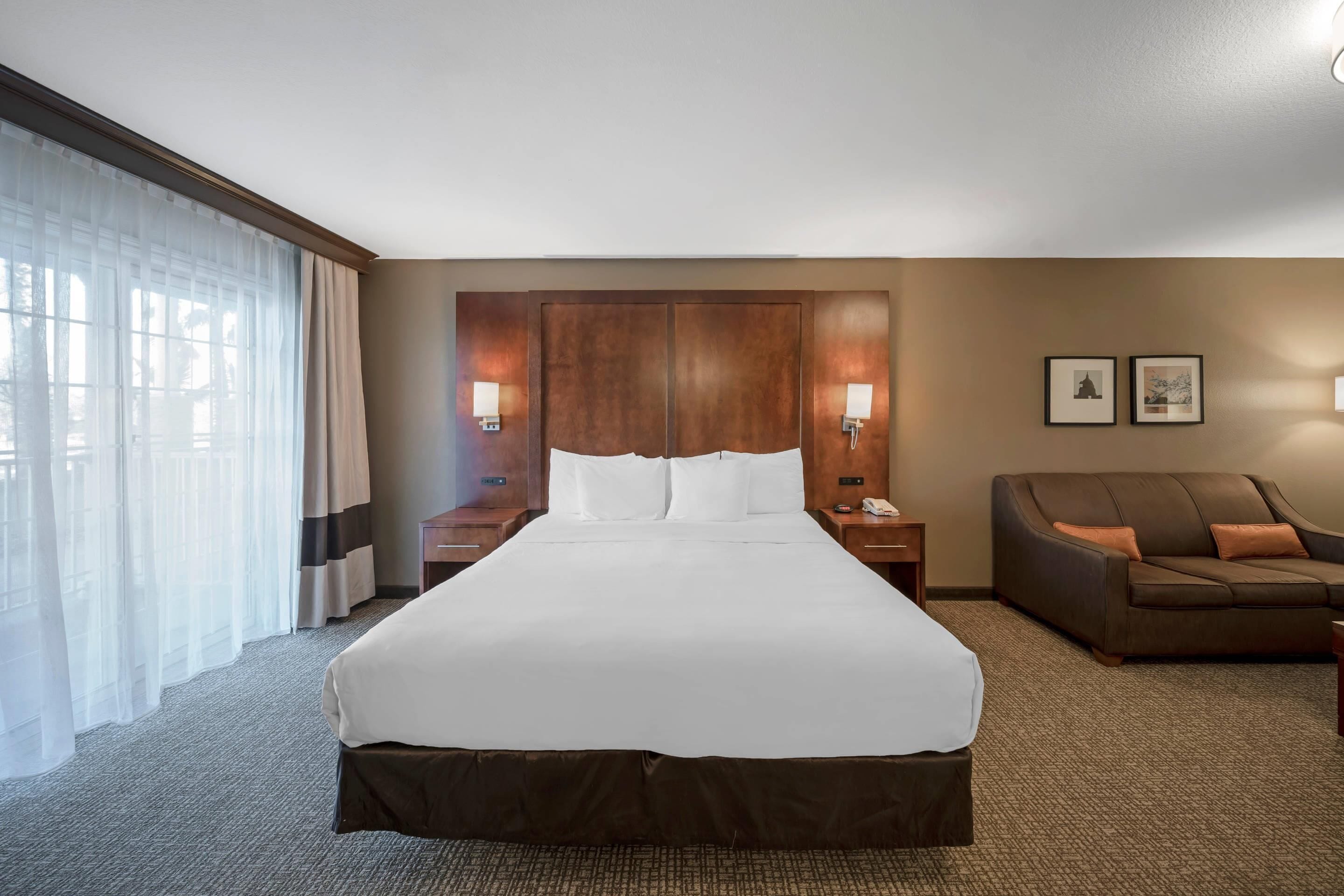 Une chambre d’hôtel avec un grand lit, une tête de lit en bois, deux tables de chevet avec des lampes, un canapé en cuir marron et une grande fenêtre avec des rideaux légers.