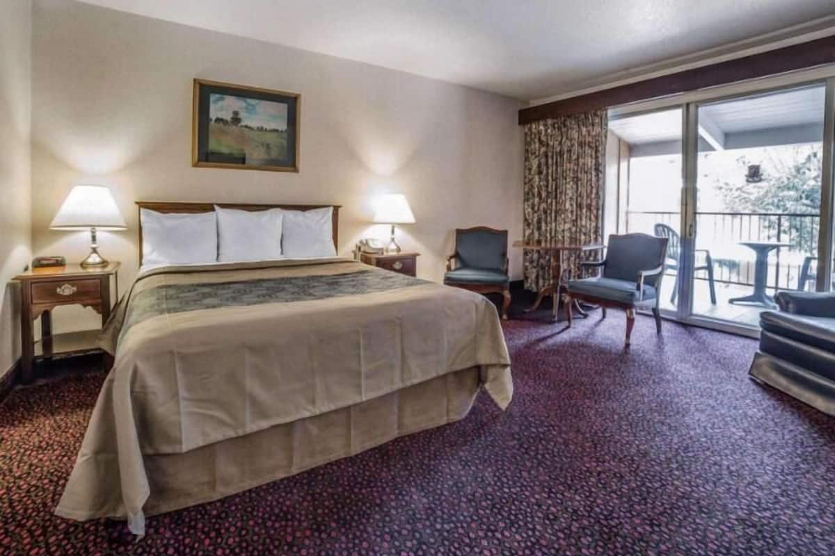 A hotel room with a bed, two bedside lamps, a chair, a small table, and a balcony with outdoor furniture.