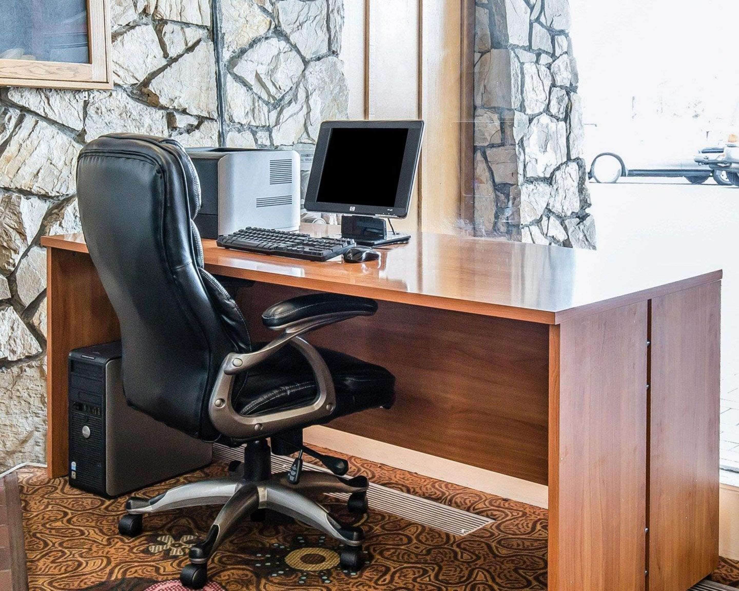A wooden desk with a computer, a black office chair, and a stone wall in the background.