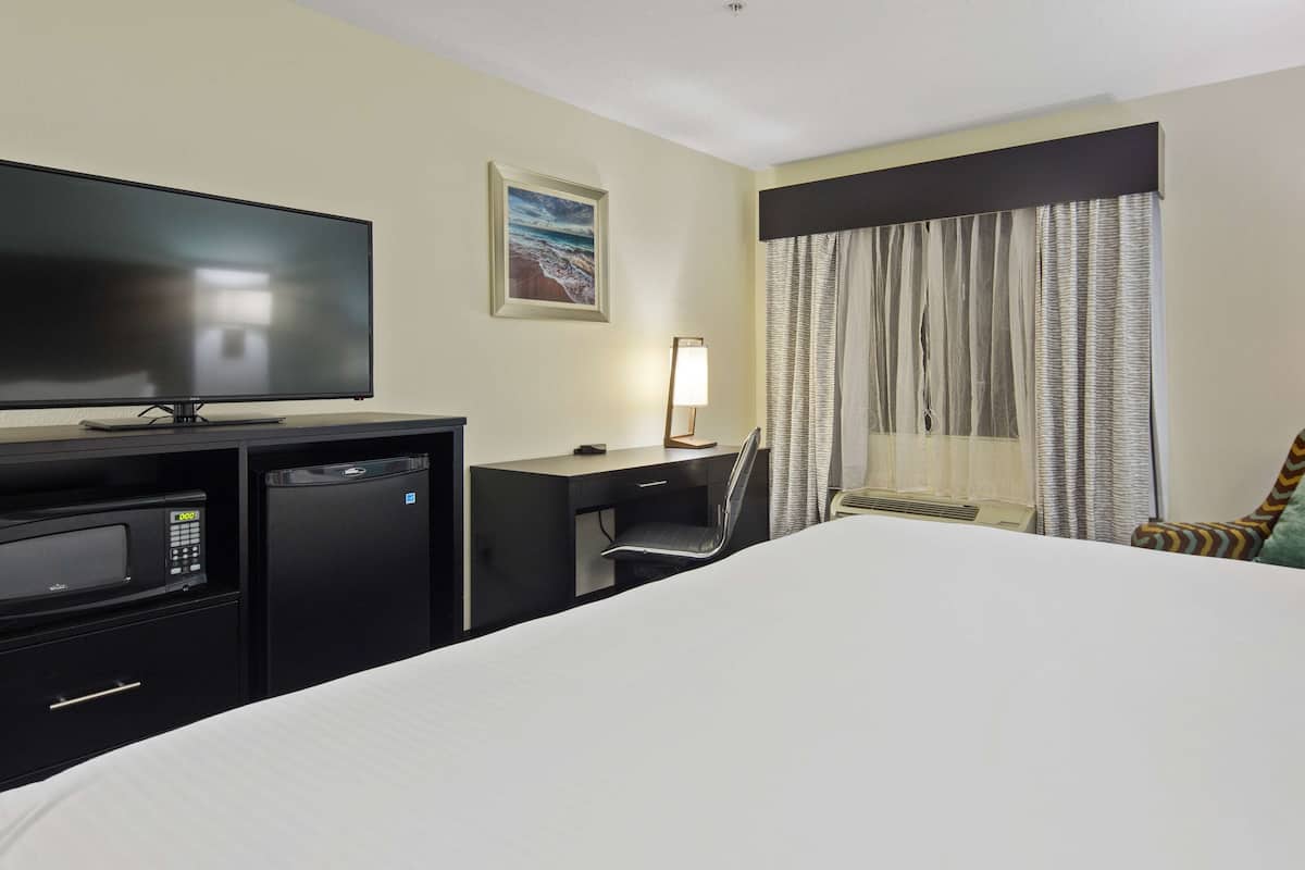 Una habitación de hotel con una cama, un escritorio con silla, televisión, refrigerador pequeño, lámpara y ventana con cortinas.