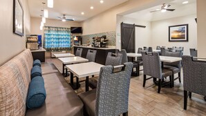 A dining area with tables and chairs, a bar area with kitchen appliances, and a seating area with a sofa and a television. - Best Western Mayport Inn & Suites (Atlantic Beach)