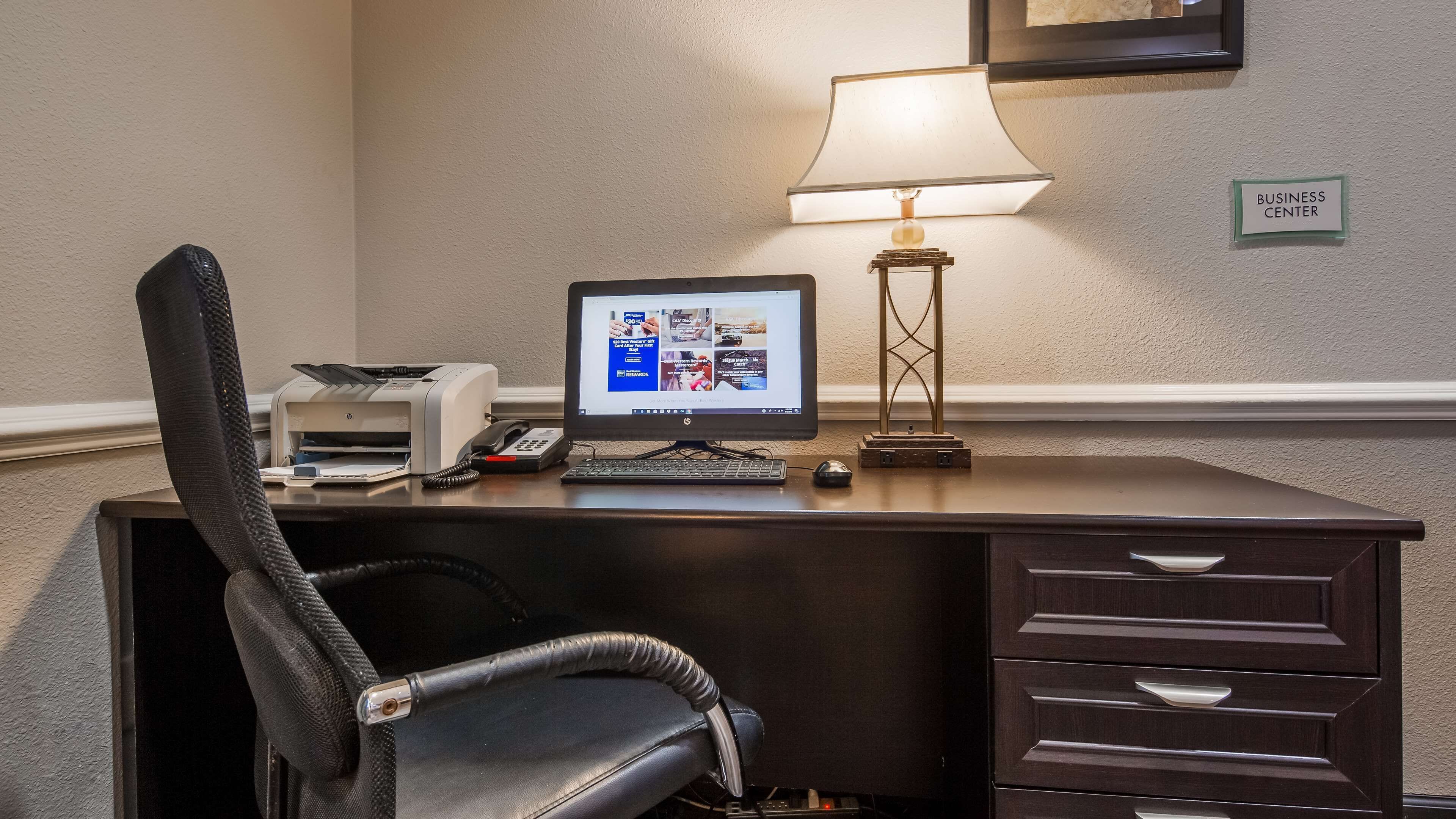A hotel business center desk with a computer, phone, printer, and lamp.
