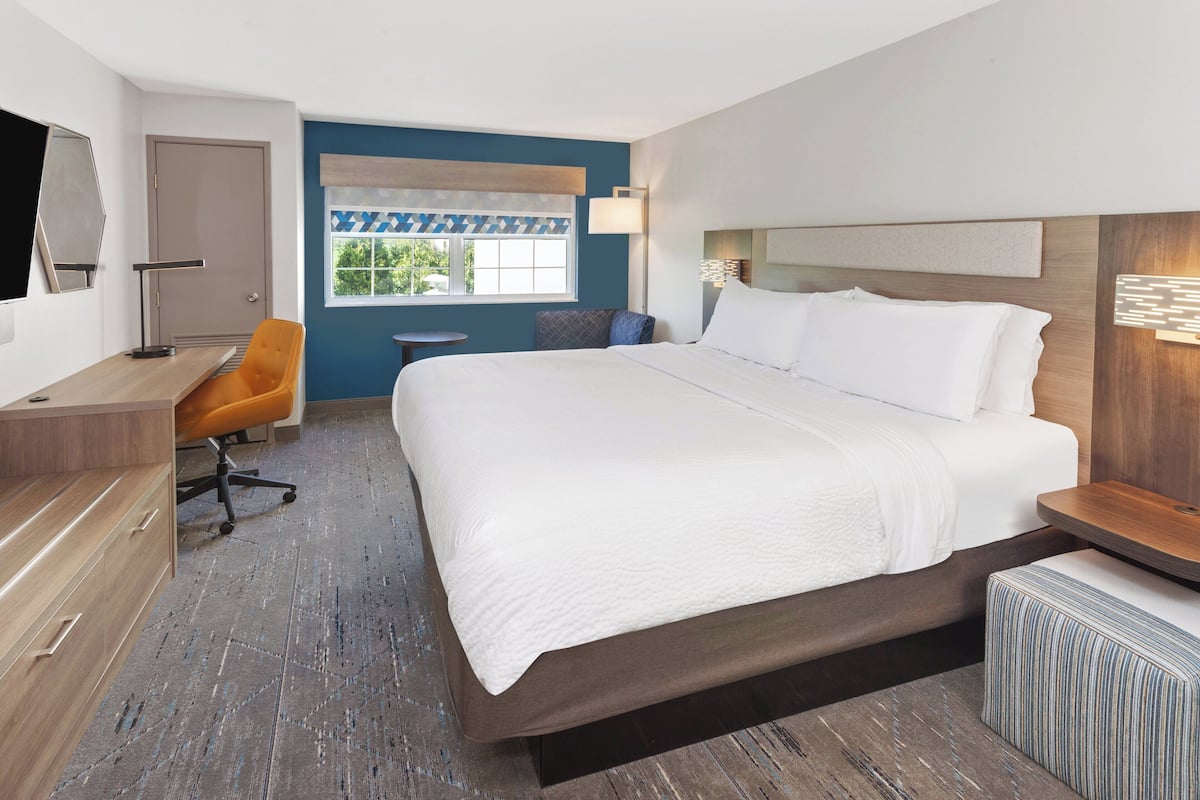 A modern hotel room with a large bed, a desk with a chair, a small round table, and a window with patterned curtains.