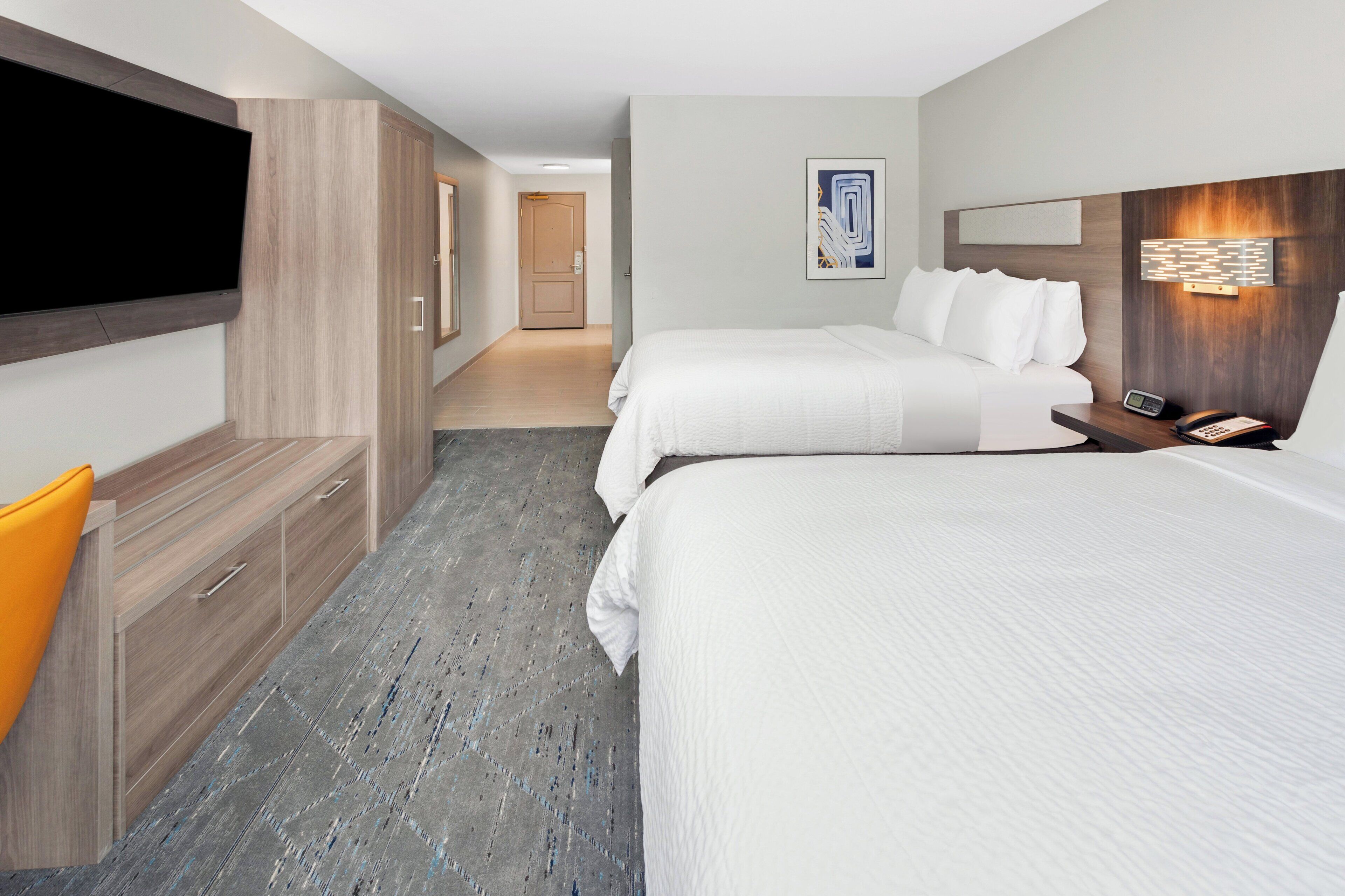 Habitación de hotel con televisión de pantalla plana en la pared, un mueble de madera con cajones, un escritorio con teléfono, dos camas con ropa blanca y un pasillo que conduce a otra habitación.
