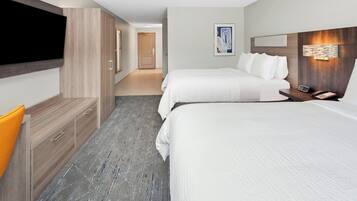 A hotel room with a flat-screen TV mounted on the wall, a wooden cabinet with drawers, a desk with a telephone, two beds with white bedding, and a hallway leading to another room.