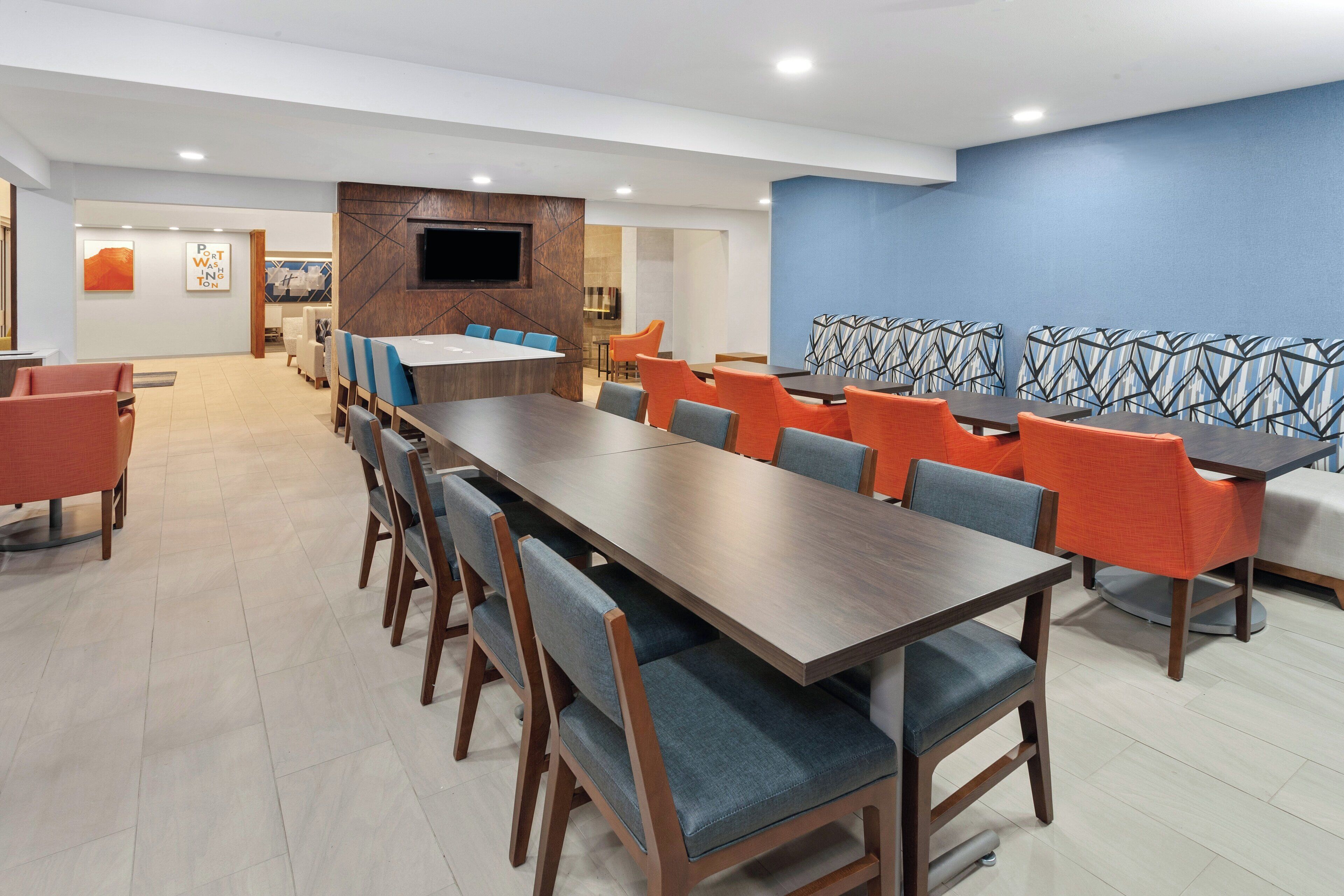 Un salón de conferencias moderno con una mesa larga, sillas azules y anaranjadas, y un televisor montado en la pared.