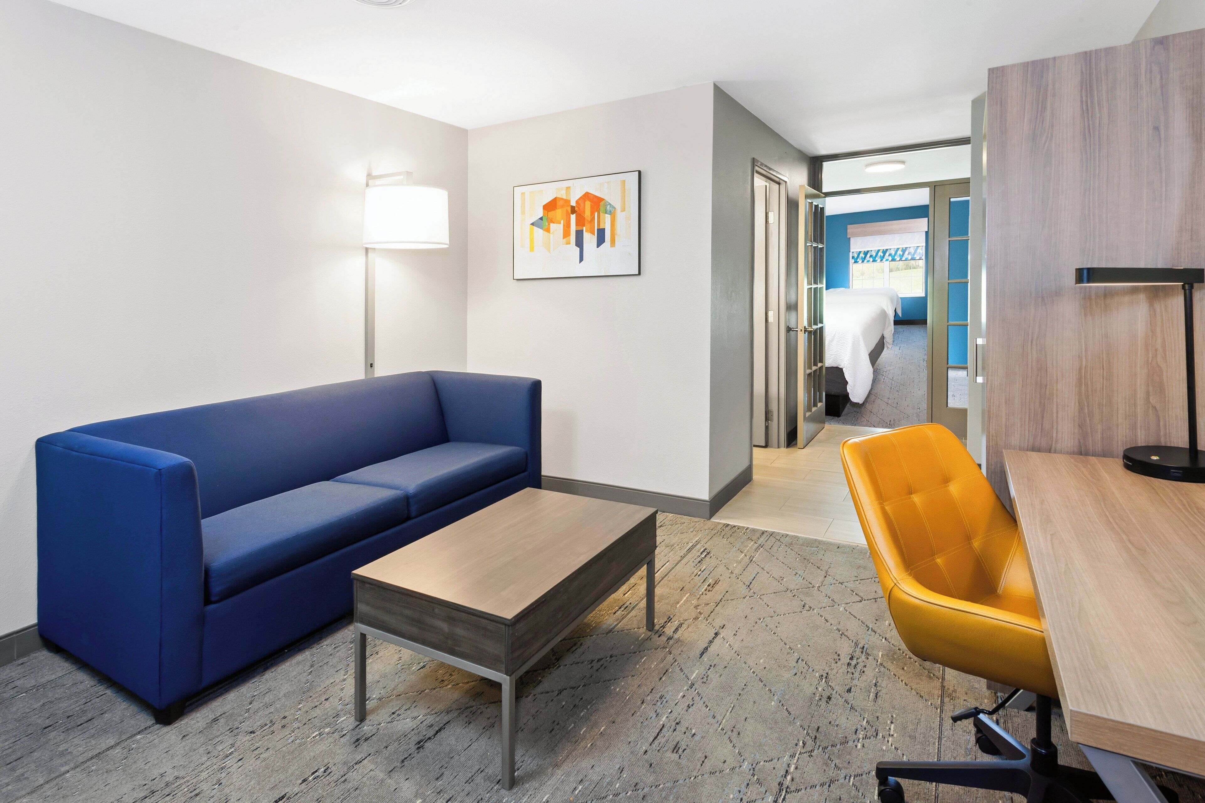 A hotel room with a blue sofa, a wooden coffee table, a yellow office chair, a wooden desk, and a bed in the background.