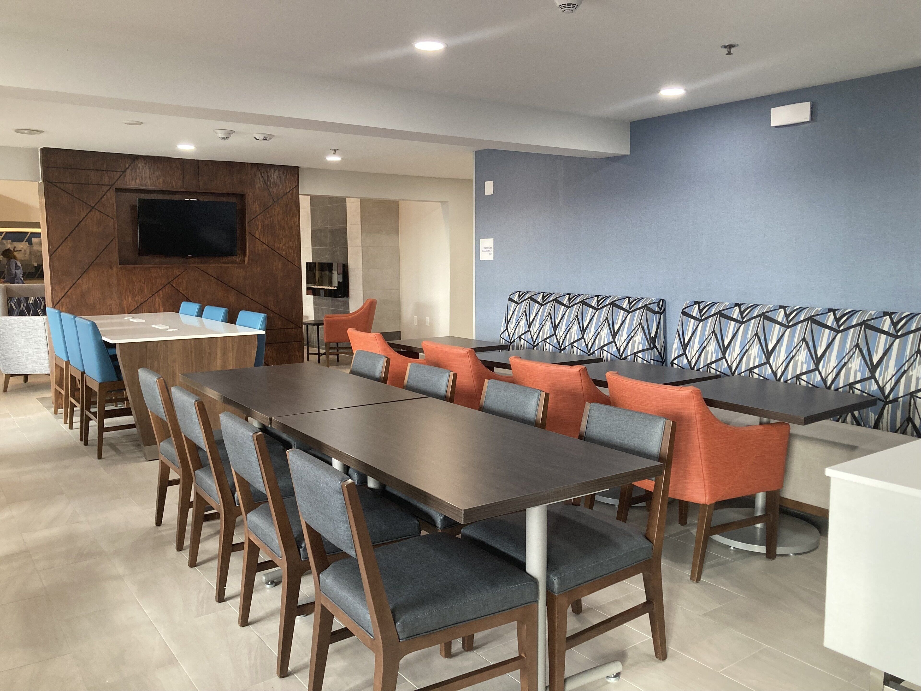 A modern dining area with a long table and chairs, a television mounted on the wall, and a patterned wall in the background.