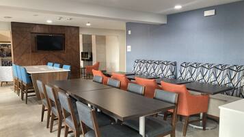 A modern dining area with a long table and chairs, a television mounted on the wall, and a patterned wall in the background.