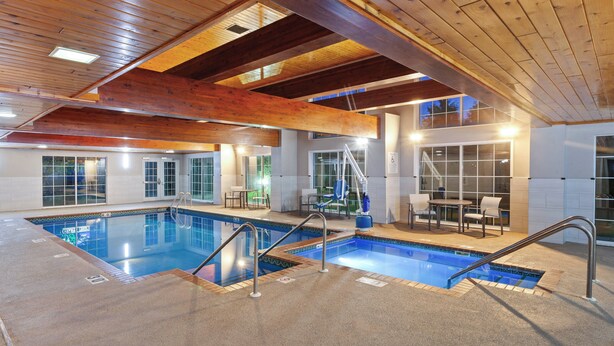 Une grande piscine intérieure divisée en deux zones distinctes, avec des poutres en bois au plafond et un espace détente comprenant des tables et des chaises.