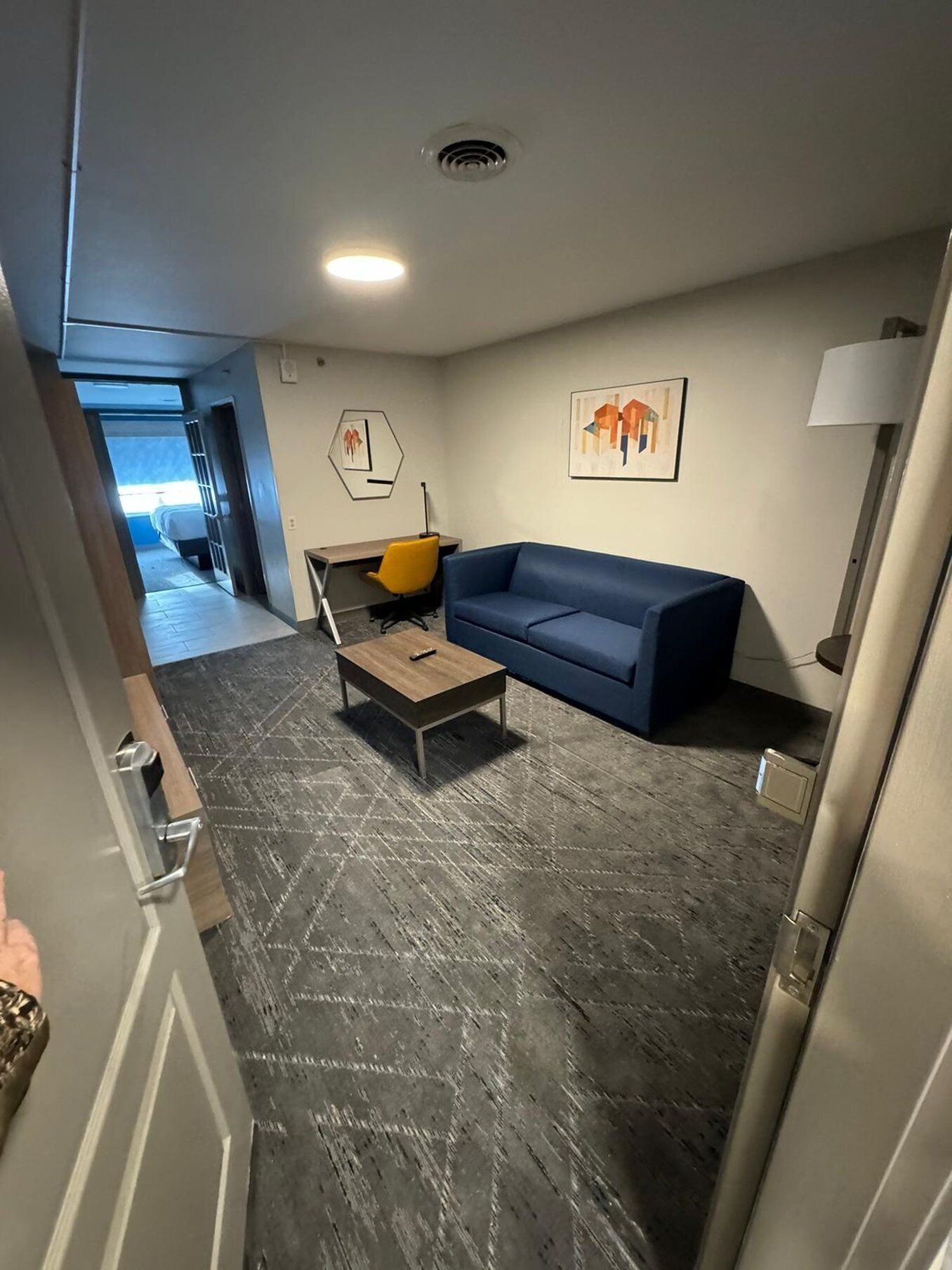 Una habitación de hotel moderna con un sofá azul, una pequeña mesa de madera y una silla amarilla. Hay un cuadro en la pared y una lámpara en la mesita de noche.