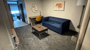 Una habitación de hotel moderna con un sofá azul, una pequeña mesa de madera y una silla amarilla. Hay un cuadro en la pared y una lámpara en la mesita de noche.