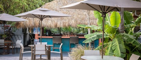 An outdoor seating area with tables and chairs, some with umbrellas, and a thatched structure in the background.