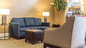 A living room with a blue sofa, a brown ottoman, and a brown armchair. There are two lamps on side tables, a large potted plant, and a bookshelf in the background. - Comfort Suites Myrtle Beach Central (Myrtle Beach)