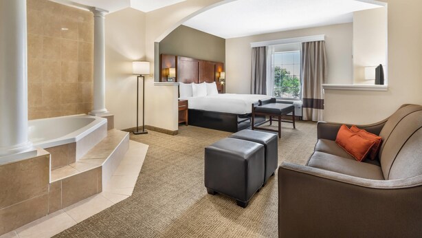 A hotel room with a large bed, a sitting area with a sofa and ottoman, a bathtub, and a window with curtains. - Comfort Suites Myrtle Beach Central (Myrtle Beach)