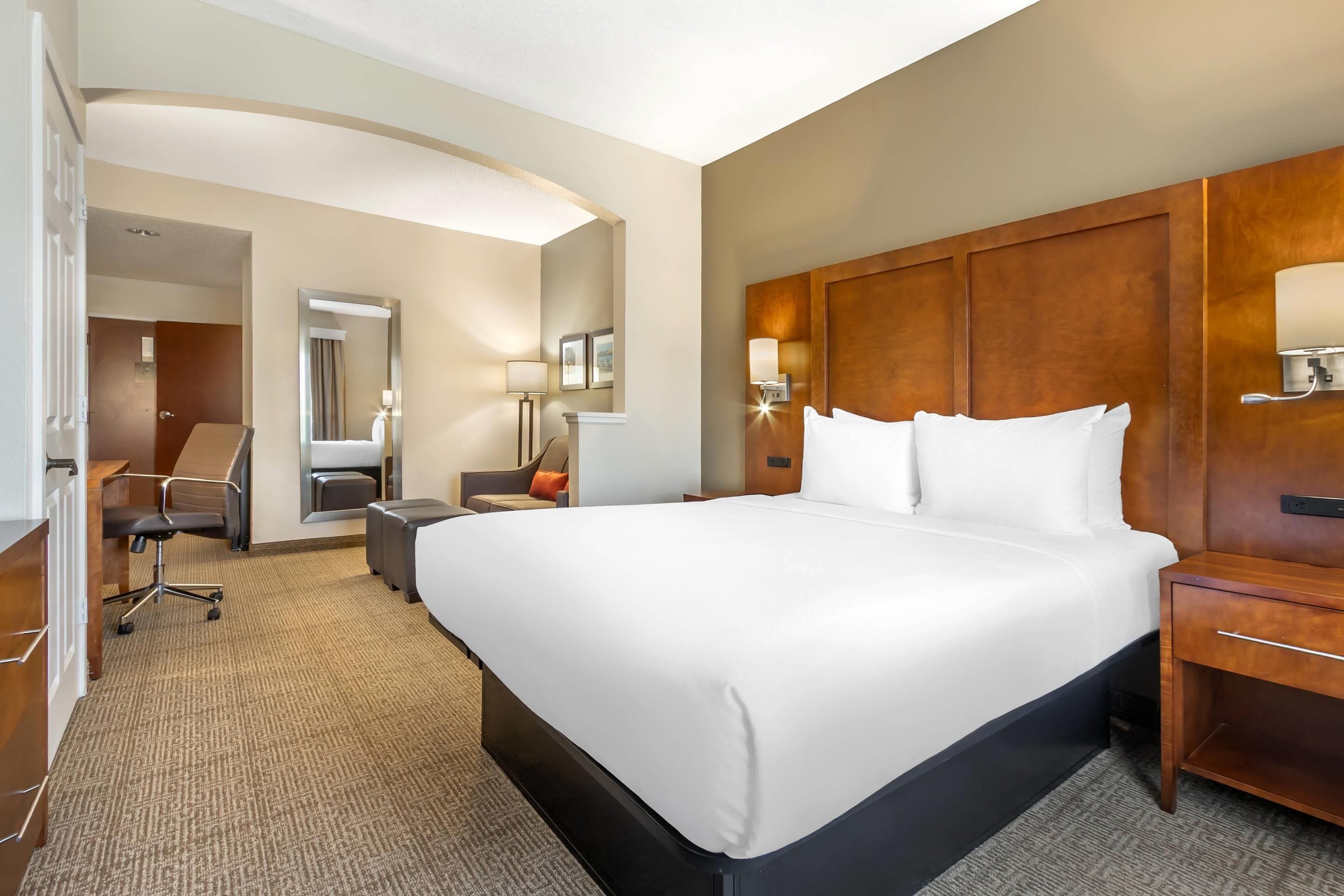 A hotel room with a large bed, a desk, a chair, a mirror, and a nightstand with a lamp.