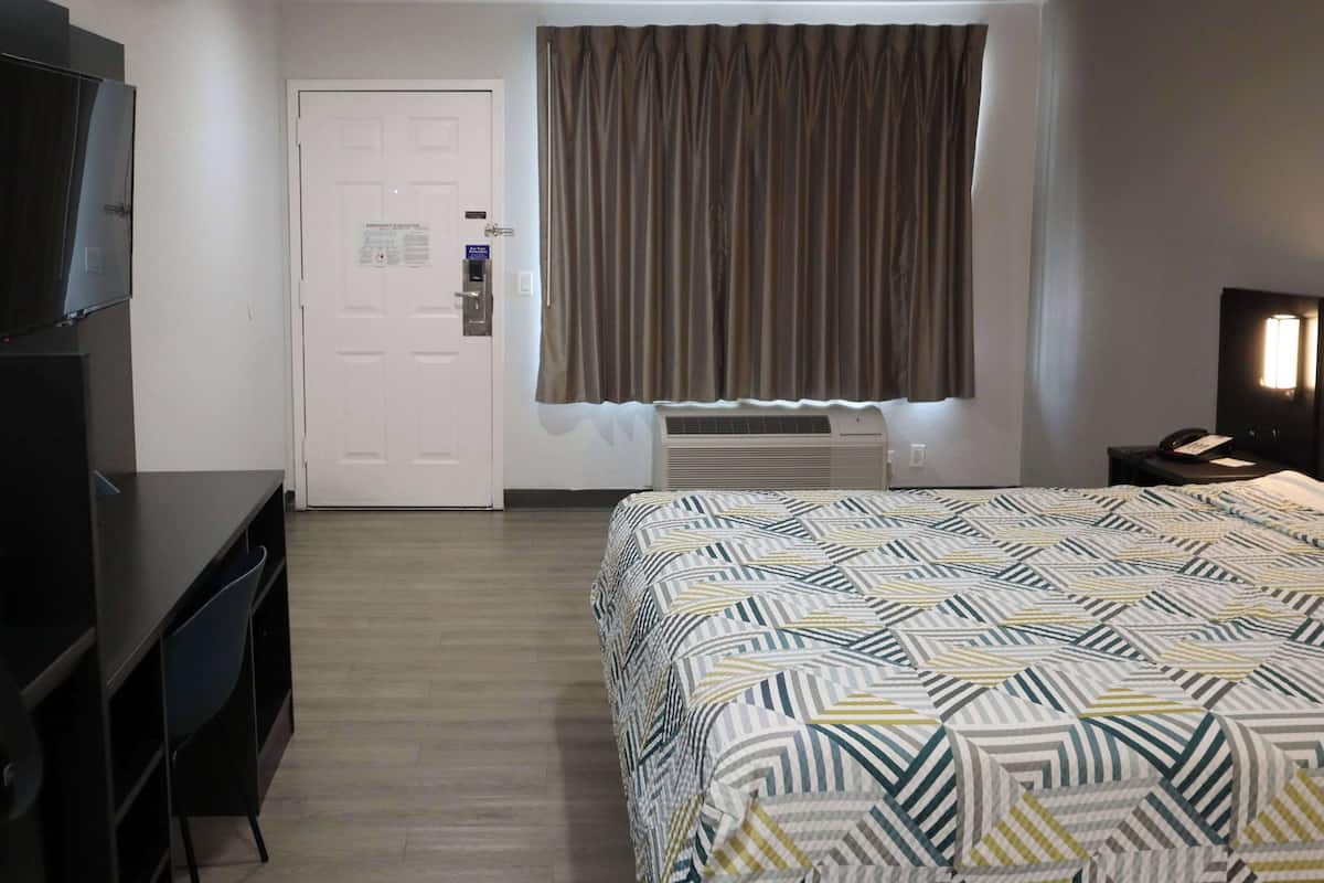 A hotel room with a bed, a desk, a TV, a window with curtains, an air conditioning unit, and a nightstand with a phone.