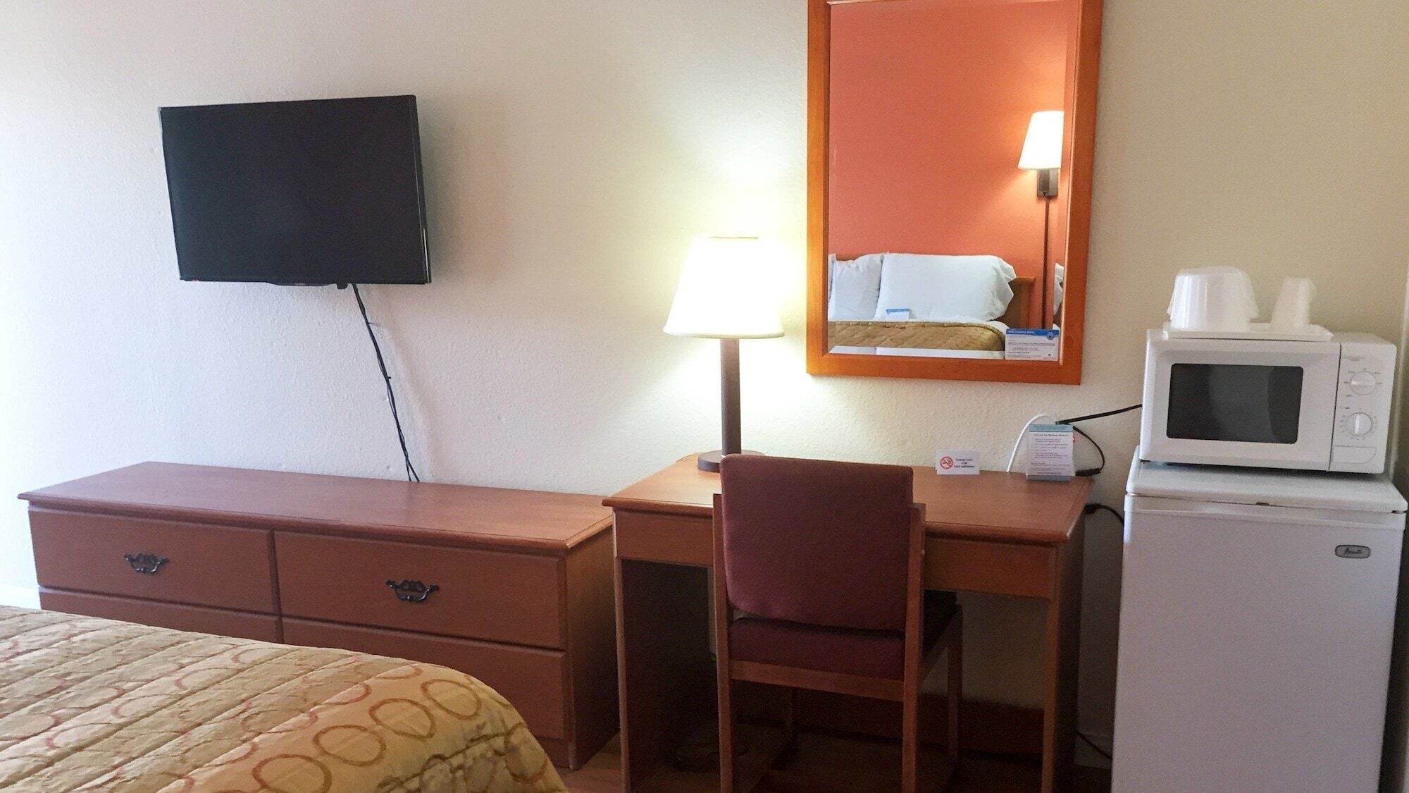 Una habitación de hotel con cama, un escritorio con silla, televisor montado en la pared, lámpara, microondas y refrigerador.