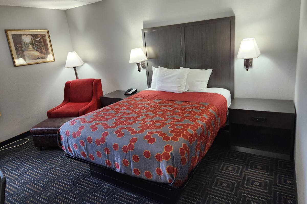 Une chambre d’hôtel avec un grand lit, deux lampes de chevet, un fauteuil rouge, un bureau avec un téléphone et un tableau encadré au mur.