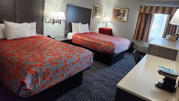 A hotel room with two beds, a desk, a chair, a lamp, a heater, and a window with curtains.