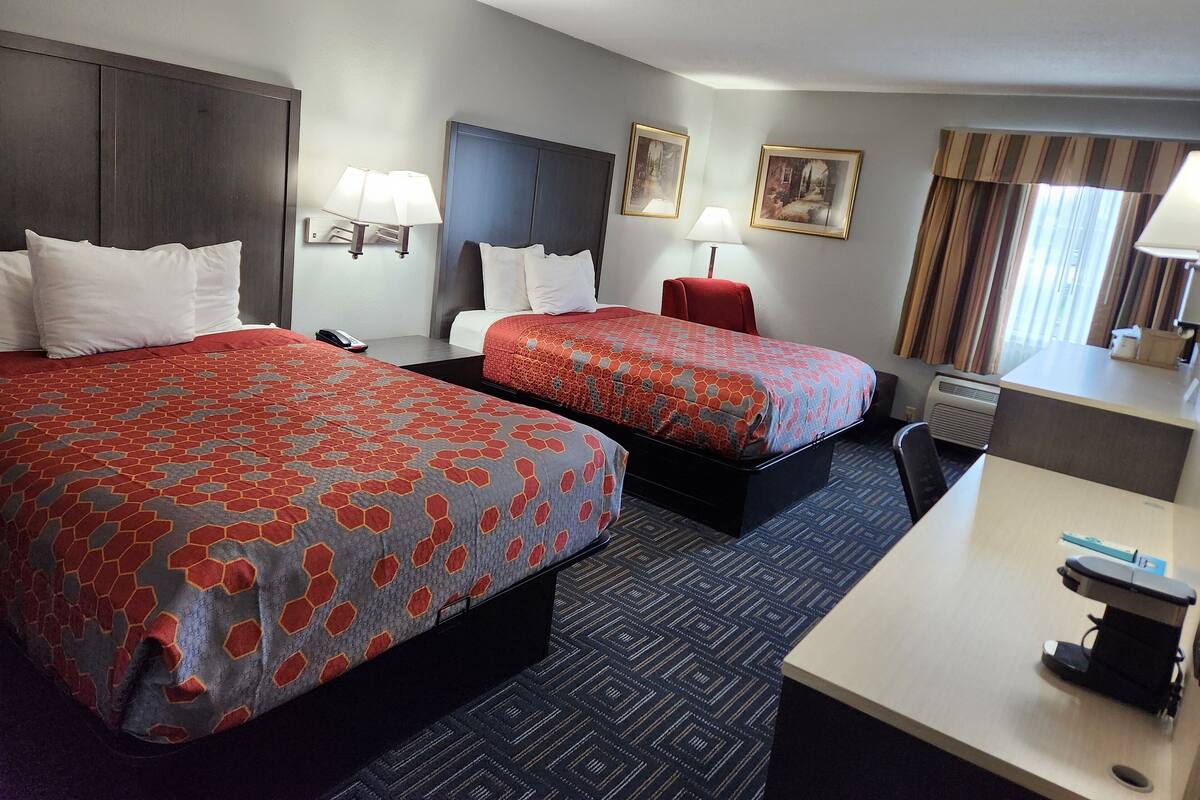 Une chambre d’hôtel avec deux lits, un bureau, une chaise, une lampe, un radiateur et une fenêtre avec des rideaux.