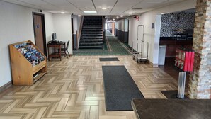 Um hall de hotel com padrão geométrico no chão em formato de losango, uma escadaria, um balcão de recepção e uma banca de revistas.