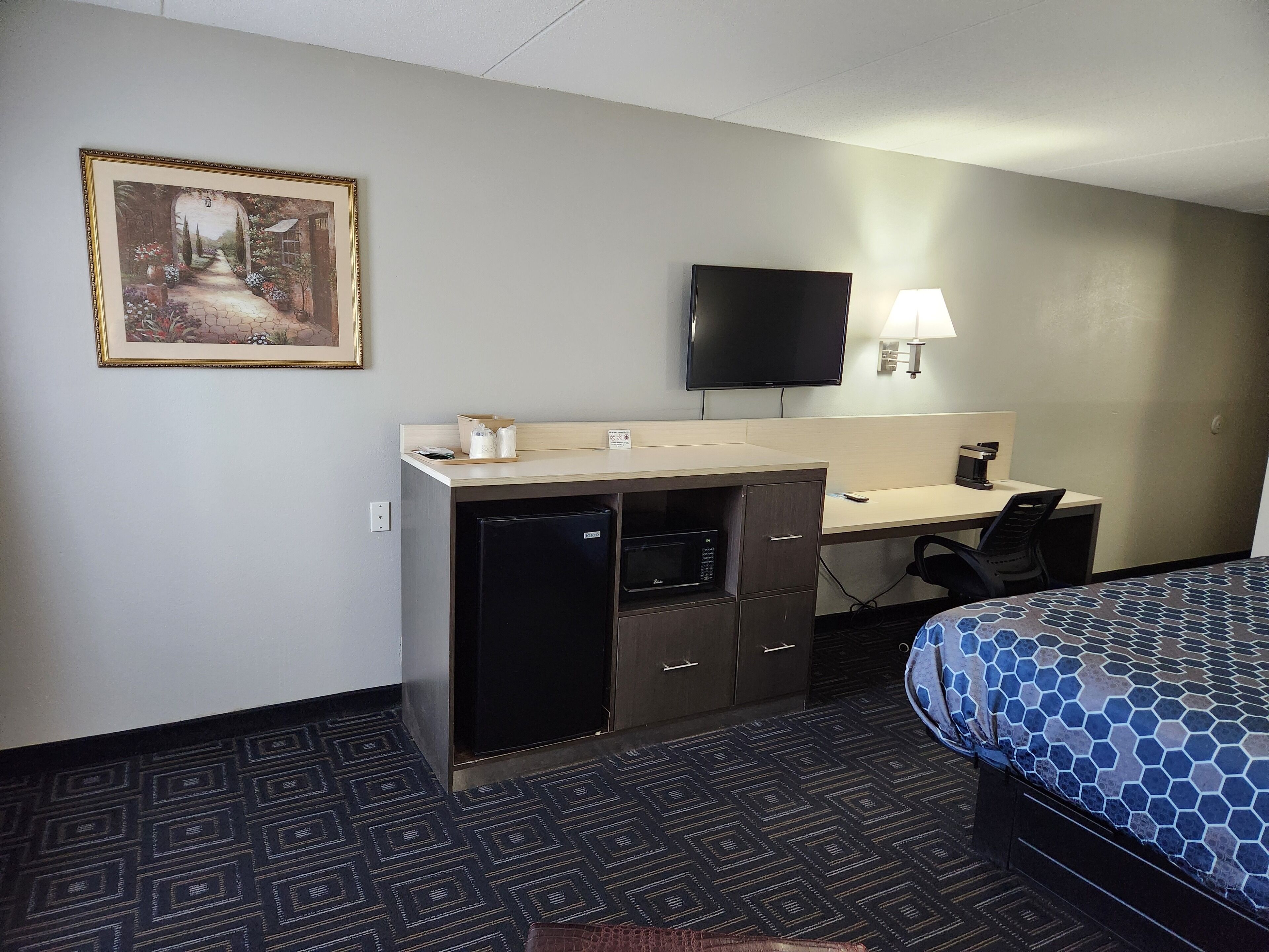A hotel room with a bed, desk, chair, television, and a painting on the wall.
