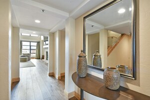 Um corredor amplo com uma mesa consola curva, dois vasos decorativos e um espelho grande que reflete a escadaria e a área de estar.