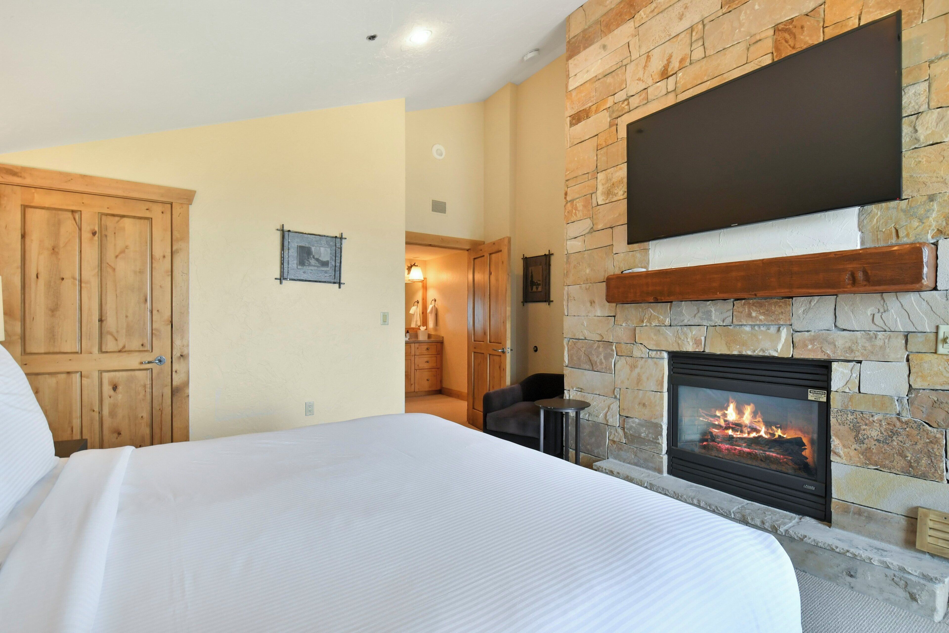 A bedroom with a large bed, a stone fireplace, a wooden door, and a mounted television.