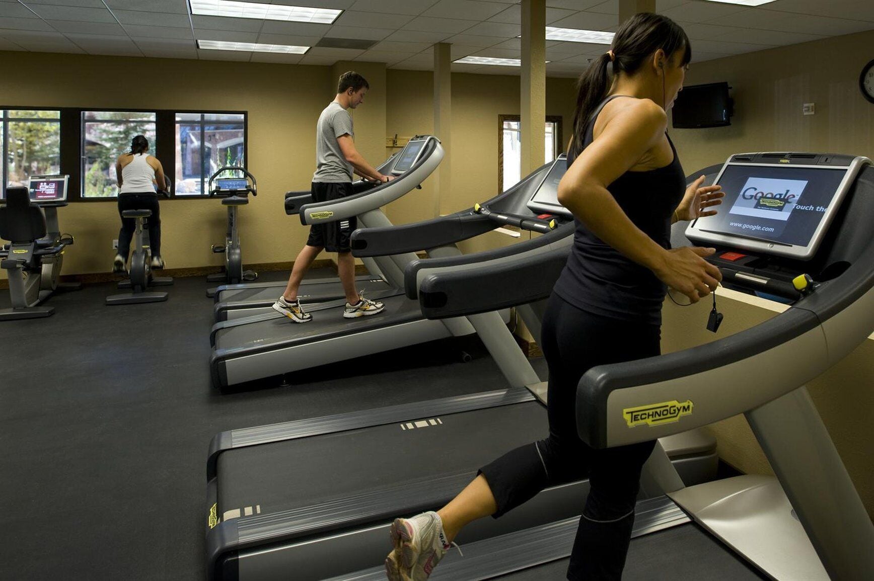 A gym with treadmills, a television, and a wall-mounted screen displaying a website.