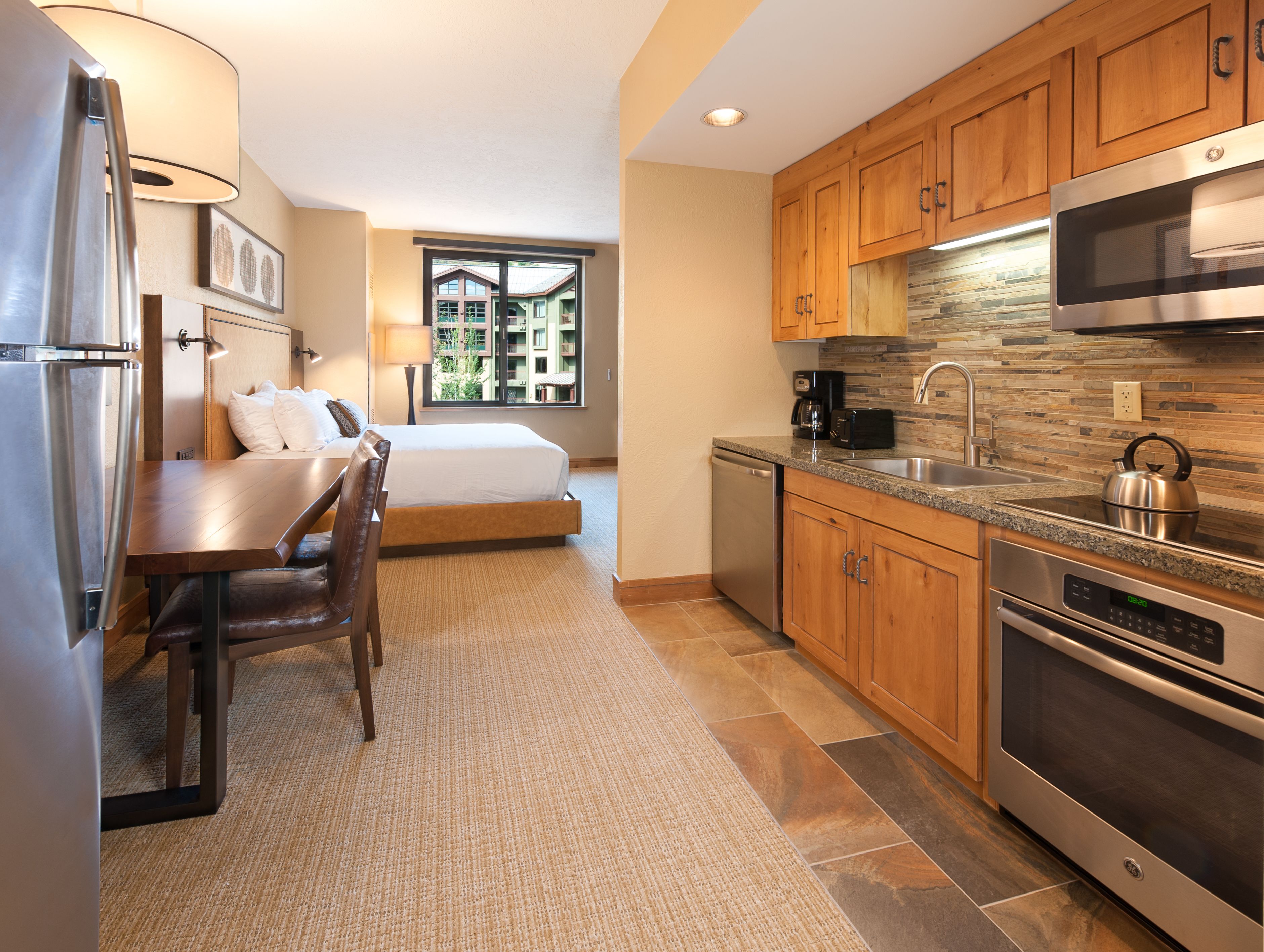 Habitación de hotel compacta con kitchenette, cama con ropa blanca, zona de comedor con mesa y sillas, y vista a un edificio a través de una ventana.