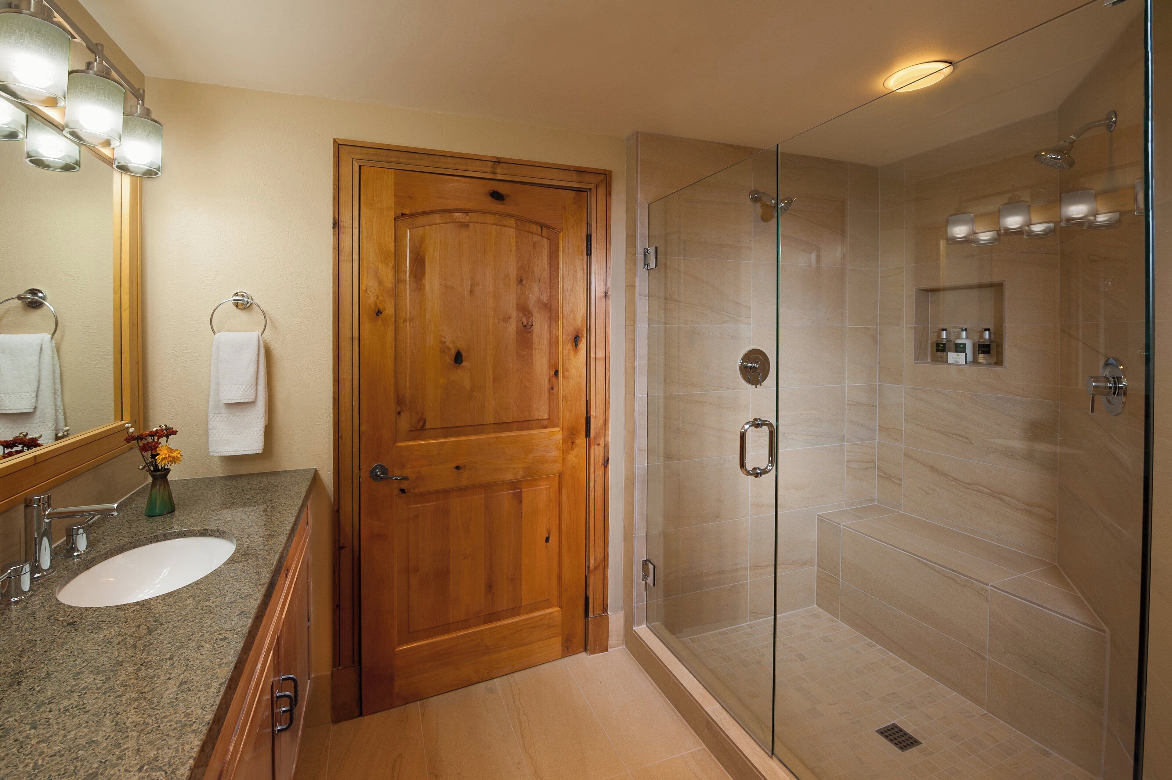 A bathroom with a wooden door, a glass-enclosed shower, and a granite countertop with a sink and mirror.