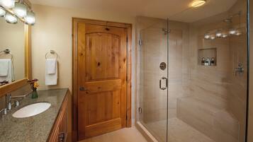 A bathroom with a wooden door, a glass-enclosed shower, and a granite countertop with a sink and mirror.