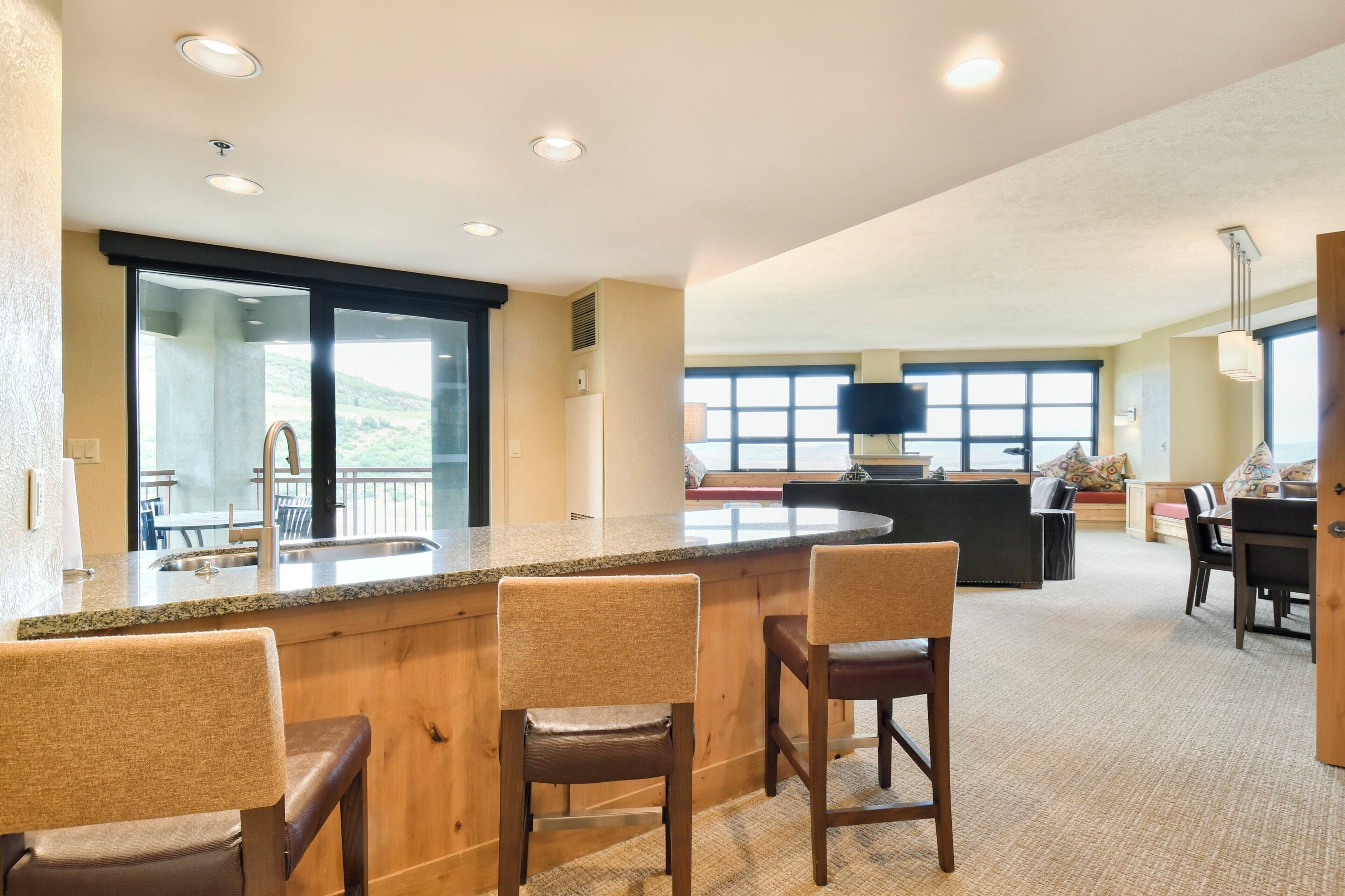 A modern kitchen with a granite countertop, wooden bar stools, and a large window overlooking a scenic view.