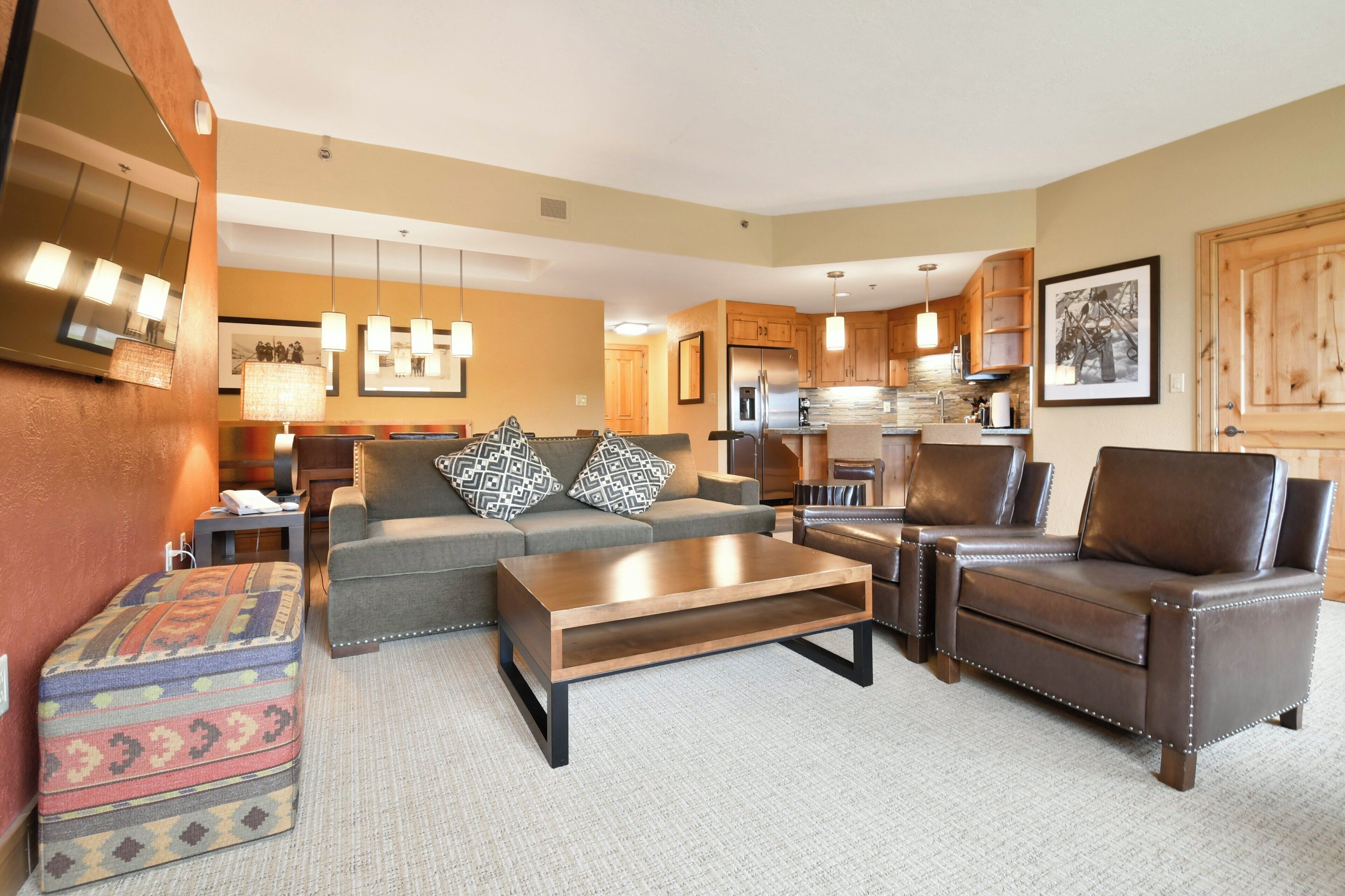 A modern living room with a sofa, coffee table, ottoman, and leather armchair. A kitchen is visible in the background.