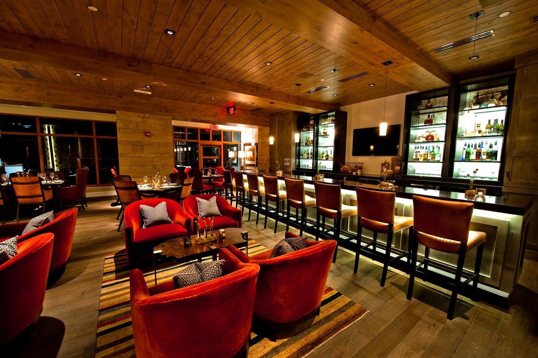 Een bar met rode bekleden stoelen, een houten plafond en een bargedeelte met flessen op de planken.