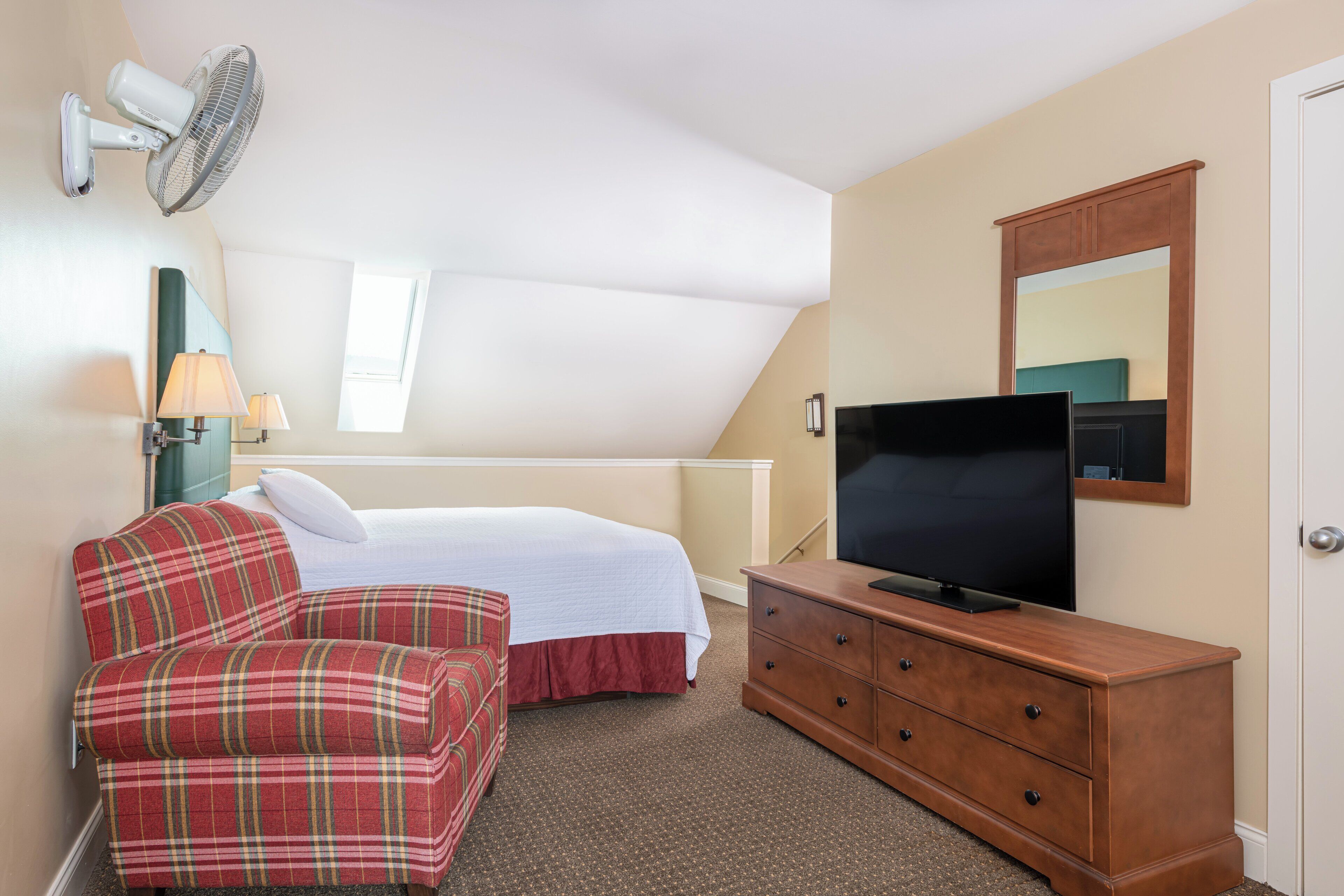 A bedroom with a bed, a plaid sofa, a dresser, a TV, a wall-mounted fan, and a window.