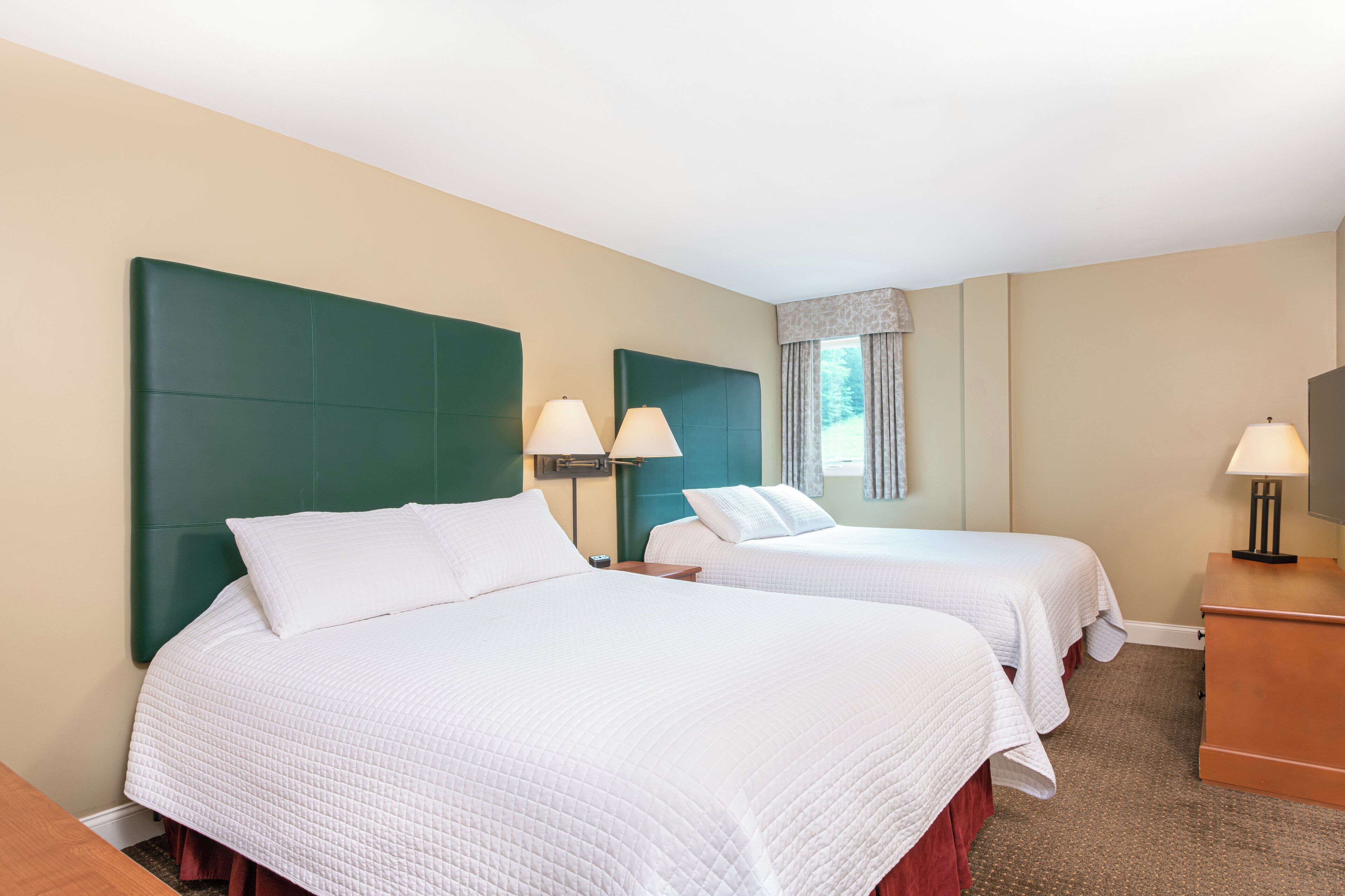 A hotel room with two beds, each with a green headboard, white bedding, and bedside lamps.