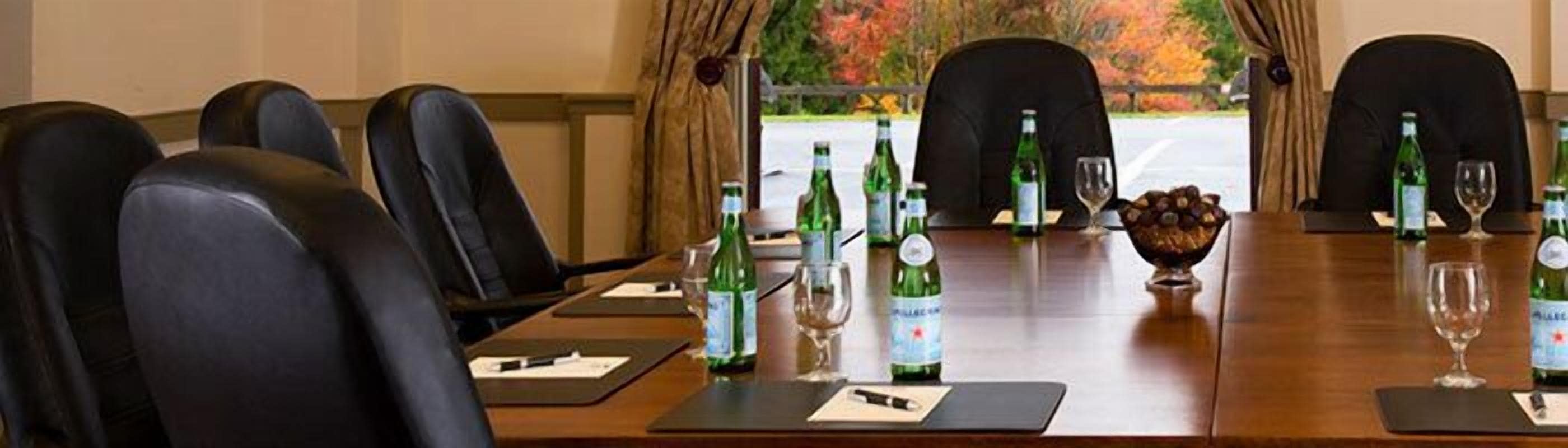 Un salón de conferencias con una larga mesa, sillas y vistas a árboles otoñales a través de un ventanal.