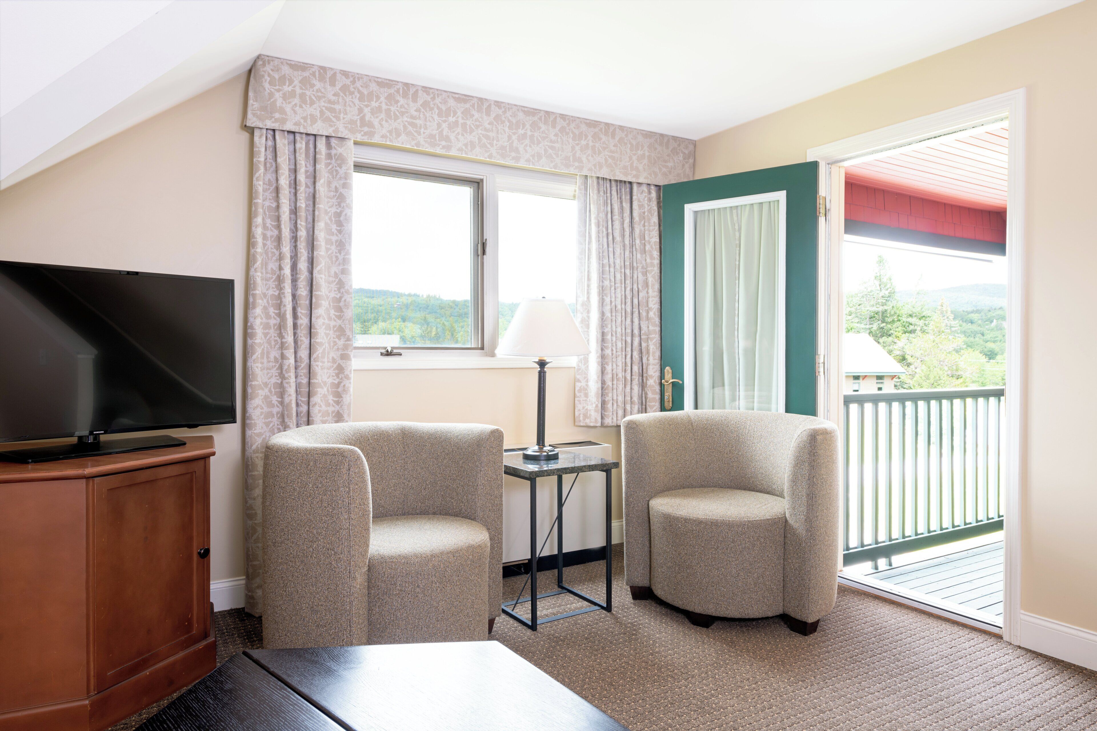 Een hotelkamer met een televisie, twee fauteuils, een tafeltje met een lamp, een raam met gordijnen en een balkon met uitzicht op de omgeving.