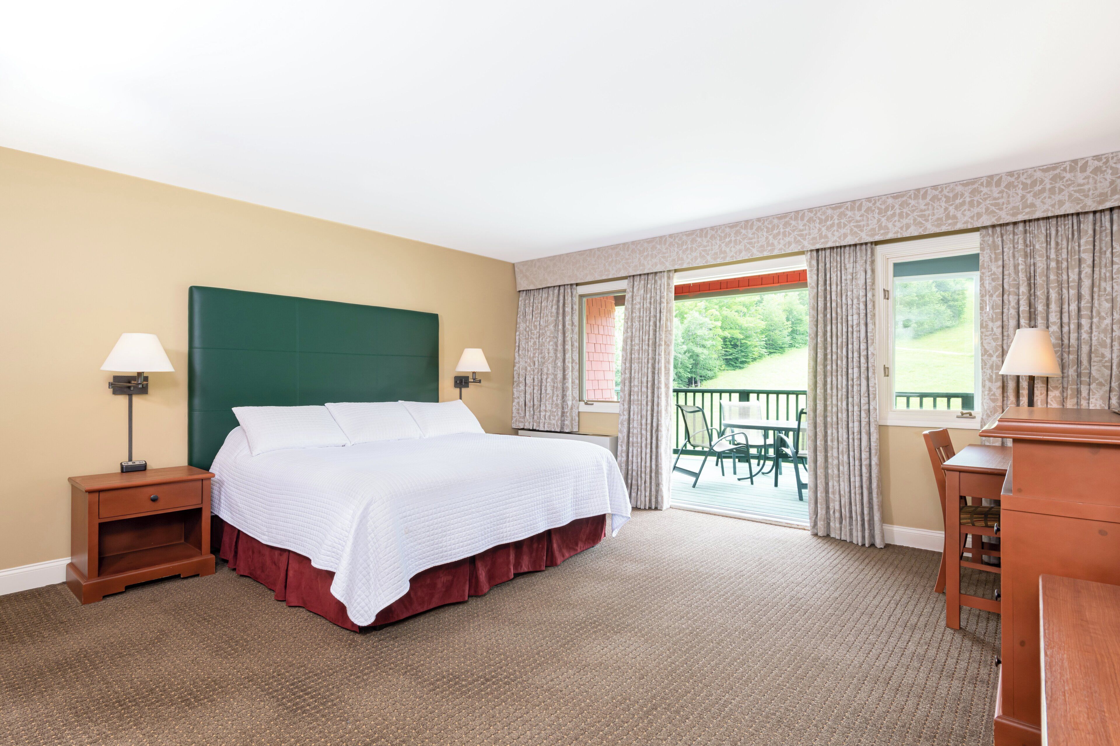 A hotel room with a large bed, two bedside tables with lamps, a wooden dresser, and a balcony with a view of greenery.