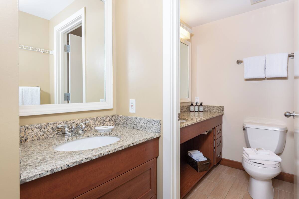 A bathroom with a granite countertop sink, a mirror, a toilet, and a vanity with drawers.