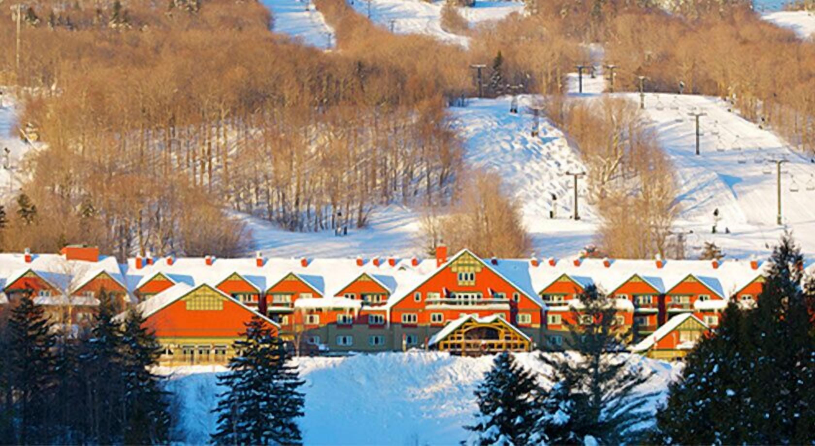 Une rangée de bâtiments aux toits rouges, recouverts de neige, probablement un hôtel ou un complexe touristique, entourés d’arbres et d’un paysage enneigé.